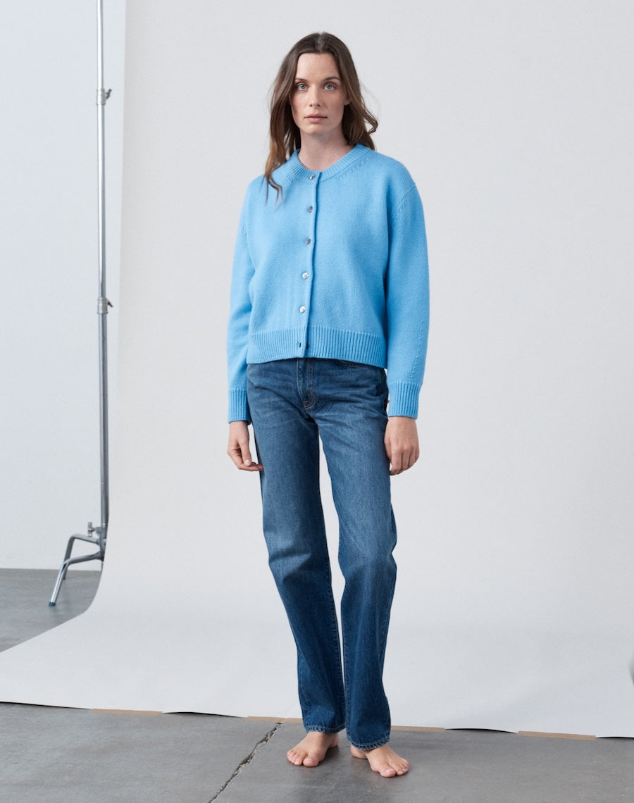 Woman wearing a light blue cardigan and dark blue jeans standing against a white background